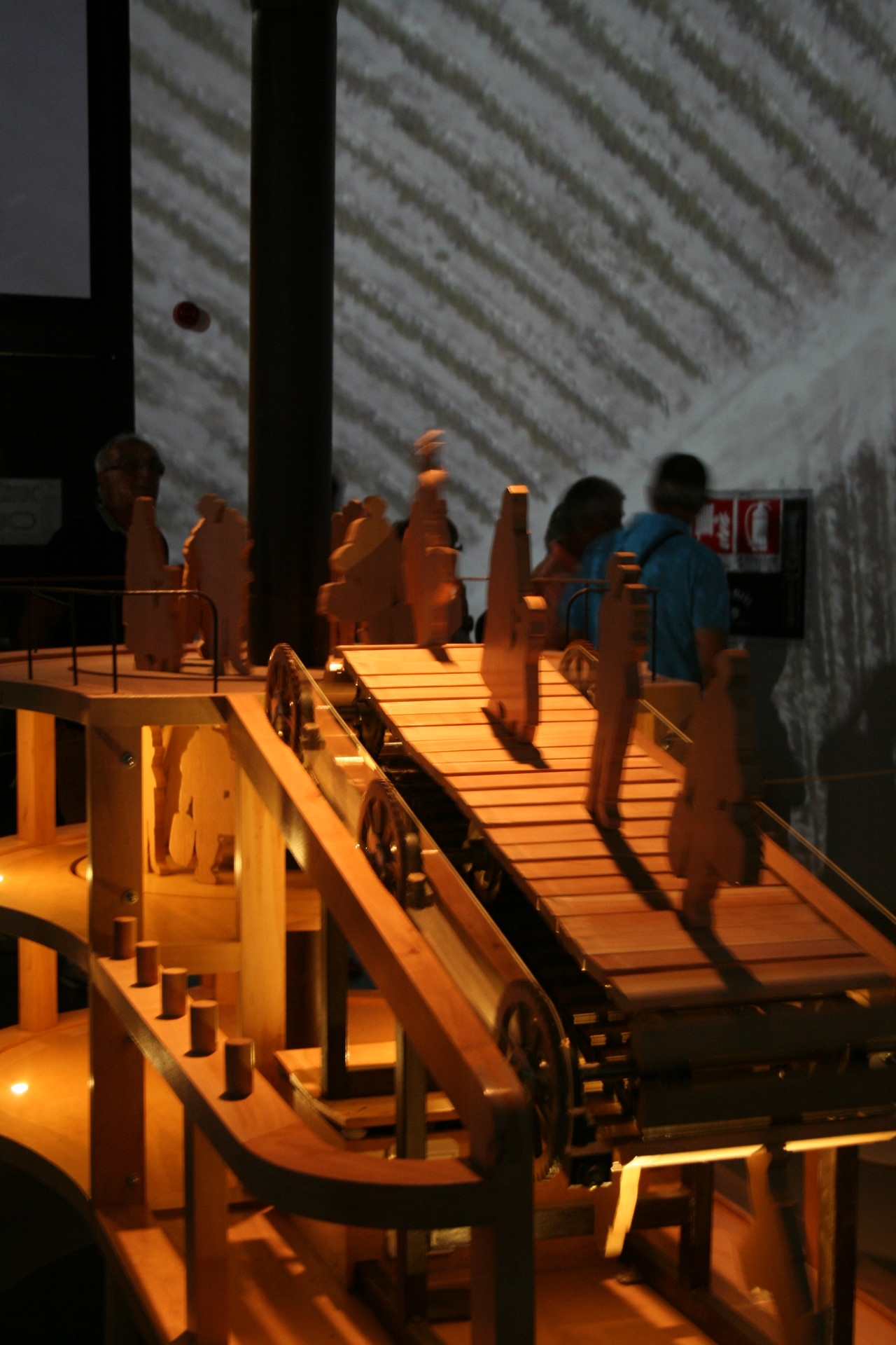 Installazione di legno raffigurante delle sagome di persone che si muovono su un nastro trasportatore anch'esso in legno